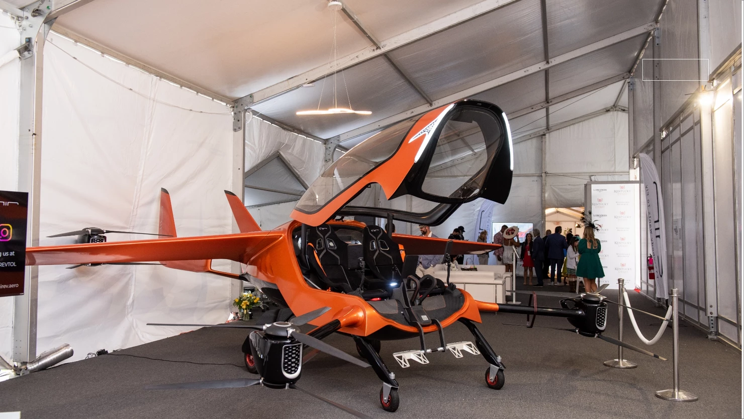An orange single-seat flying car is showcased in an exhibition tent with people in the background. The vehicle has its cockpit canopy open.