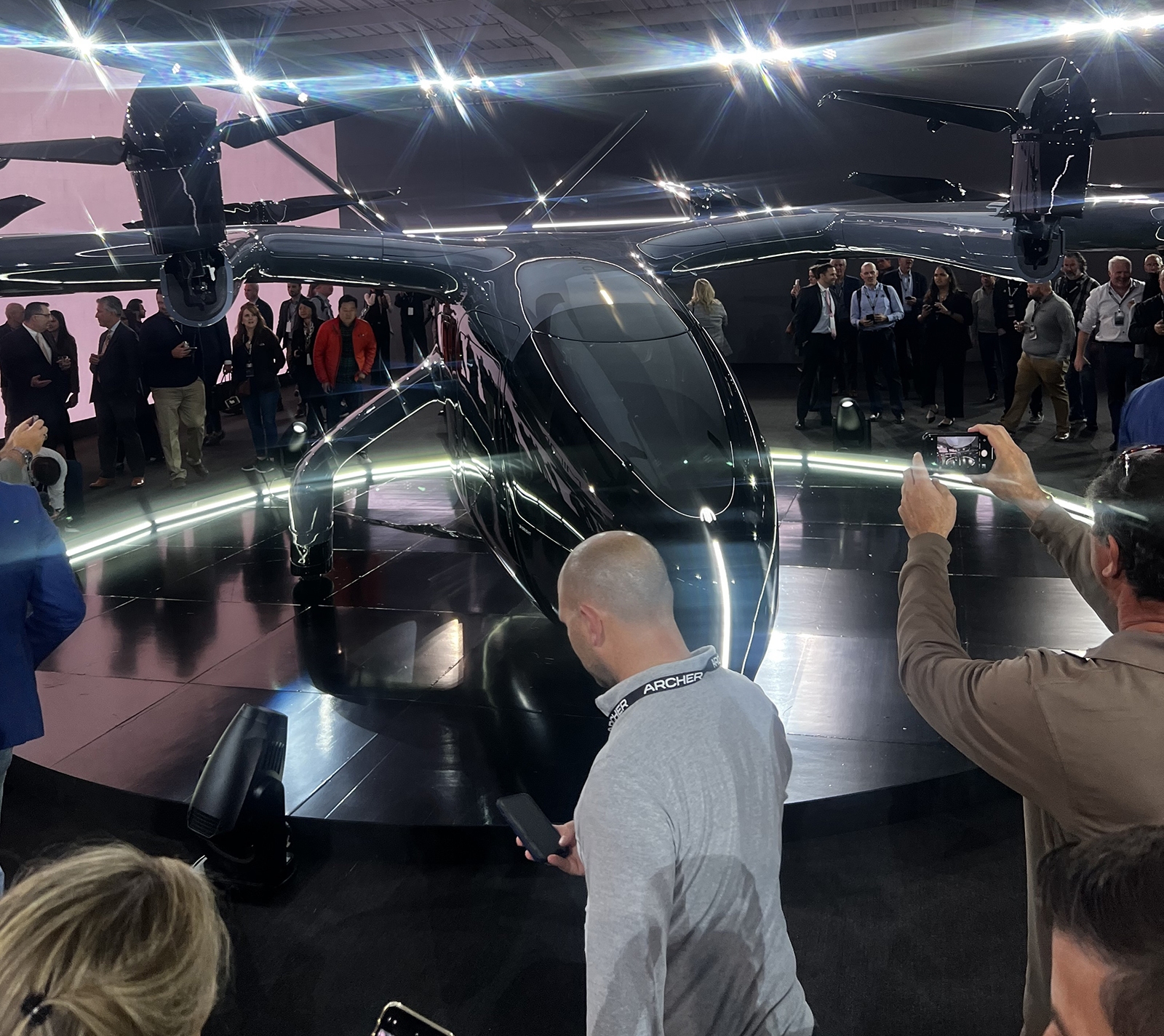 A sleek, black aircraft on display at an event, surrounded by people taking photos and observing. The aircraft is showcased on an illuminated platform.