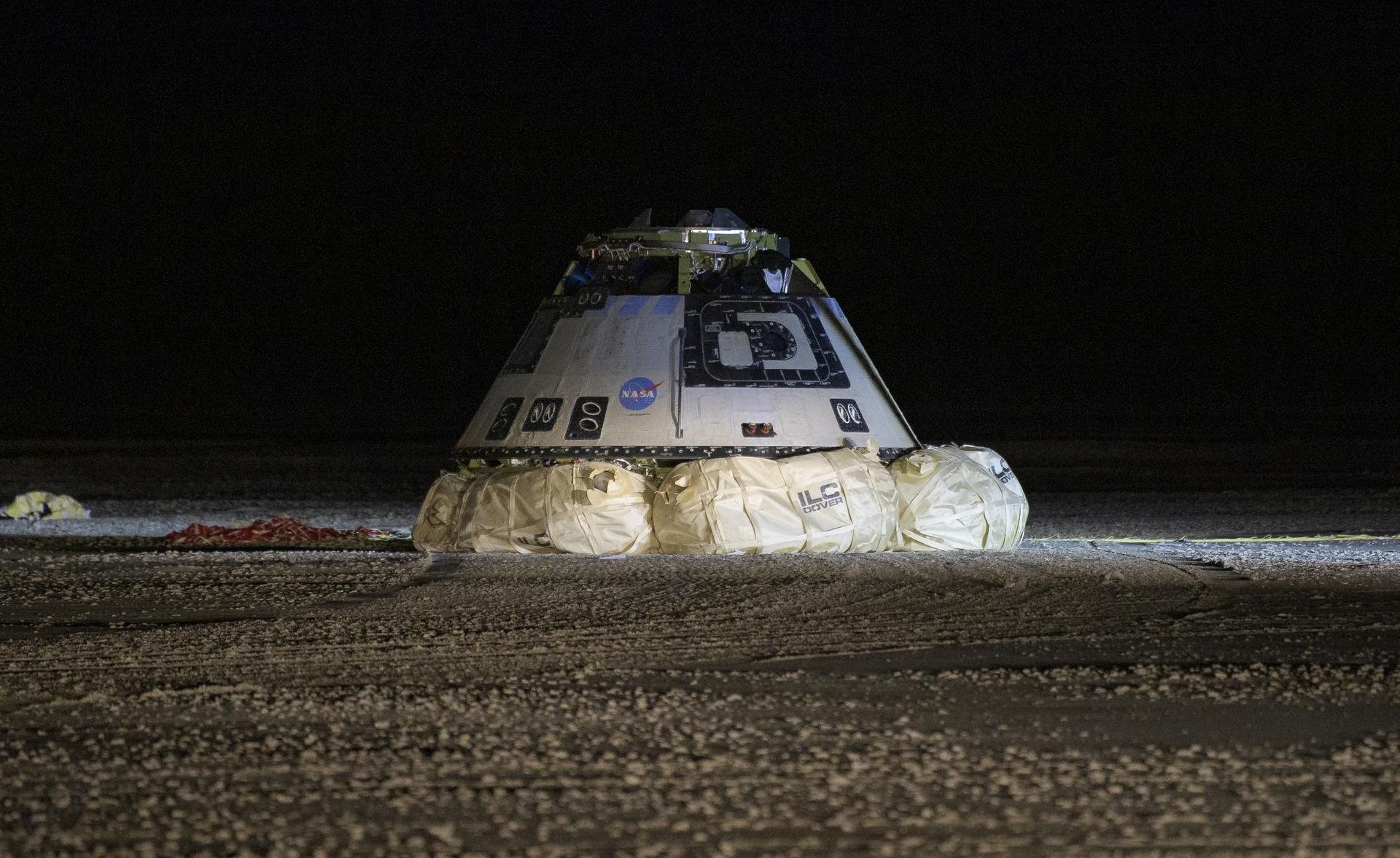 A space capsule with a NASA logo is grounded in a dark, barren landscape, supported by inflated landing airbags.