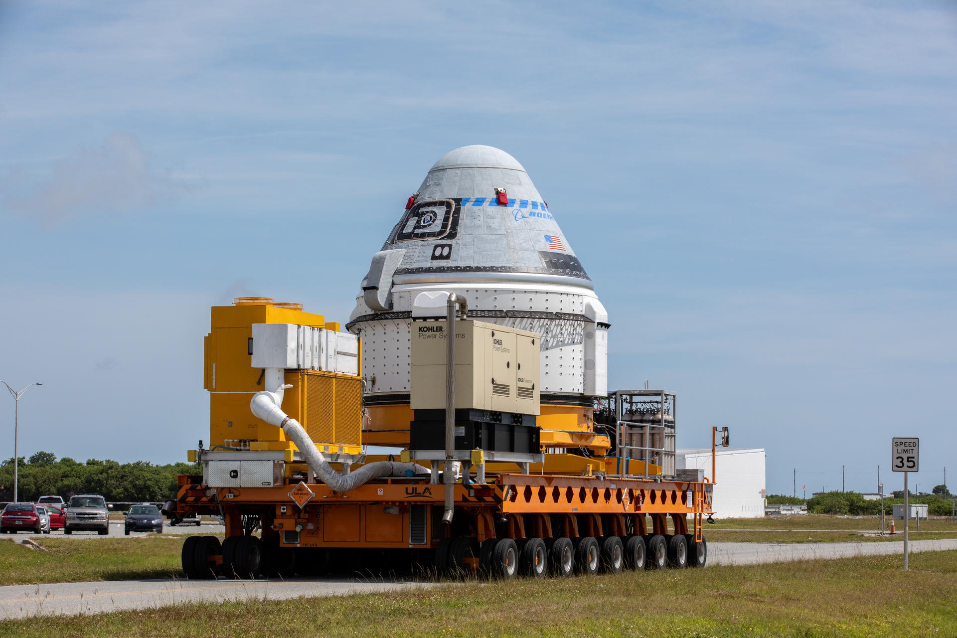 A spacecraft is being transported on a large, yellow flatbed vehicle moving along a road, with a speed limit sign of 35 mph visible to the right. There are vehicles and buildings in the background.