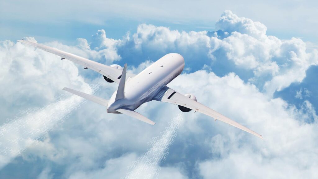 A white airplane flies above the clouds, leaving contrails in the sky. The sky is mostly cloudy with patches of clear blue.