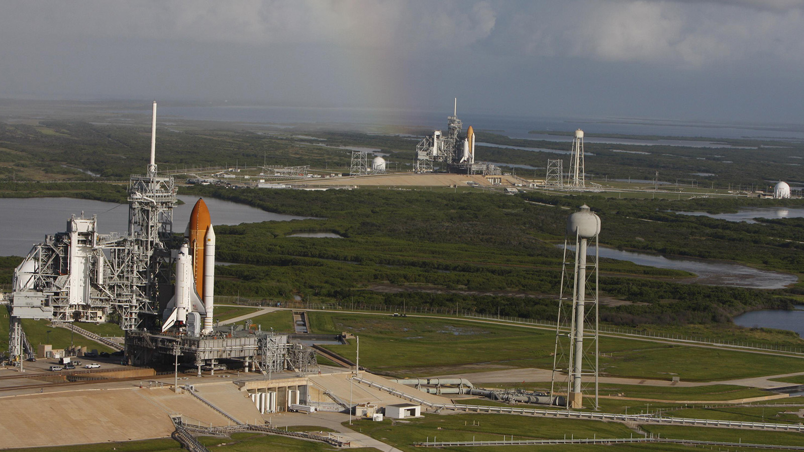 Aerial view of two space shuttles on launch pads at a space center, with a rainbow visible in the cloudy sky above. The surrounding area is green with bodies of water and infrastructure.