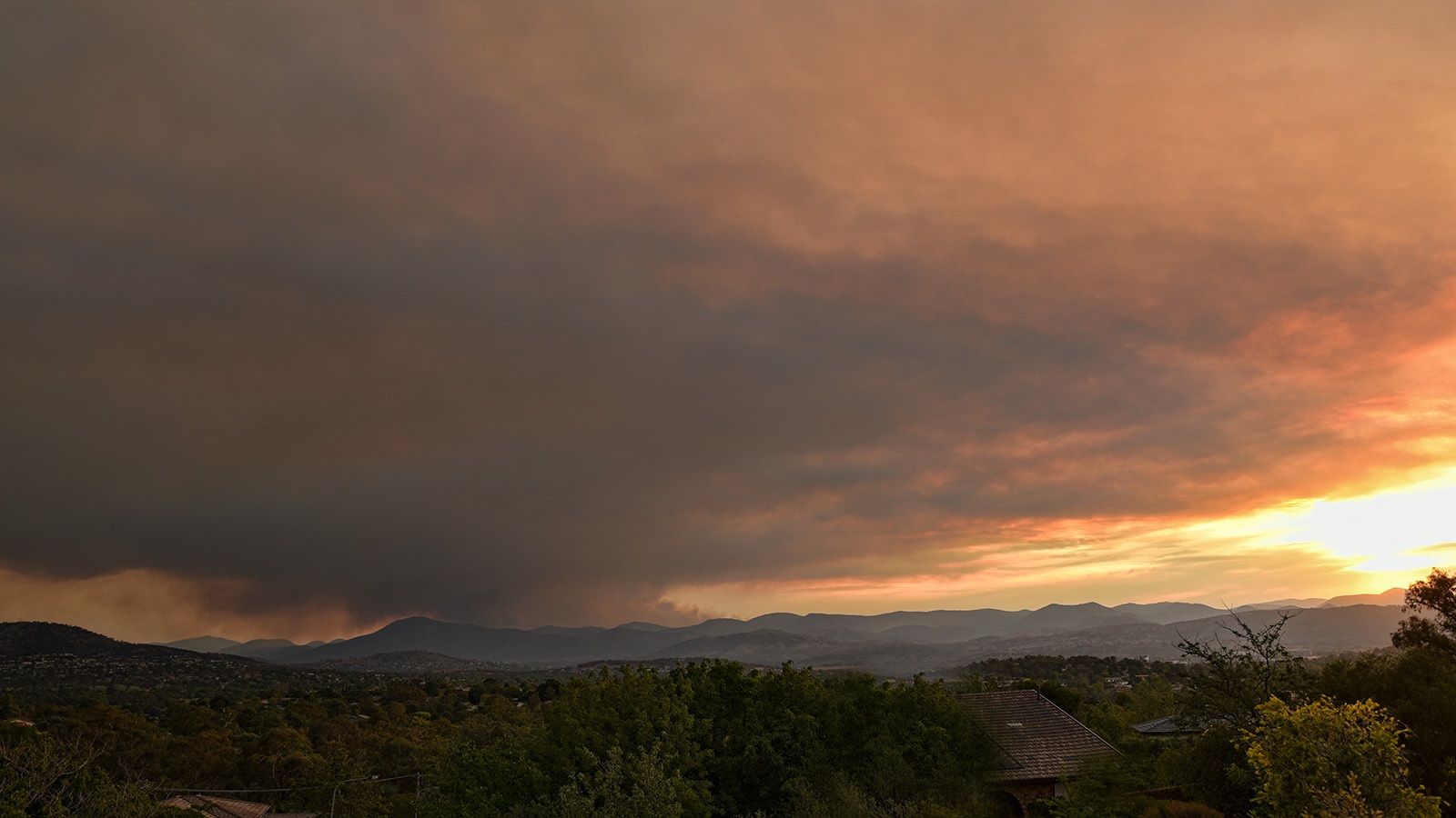 A large plume of smoke rises into the sky over a mountain range during sunset, with a landscape of trees and houses in the foreground.