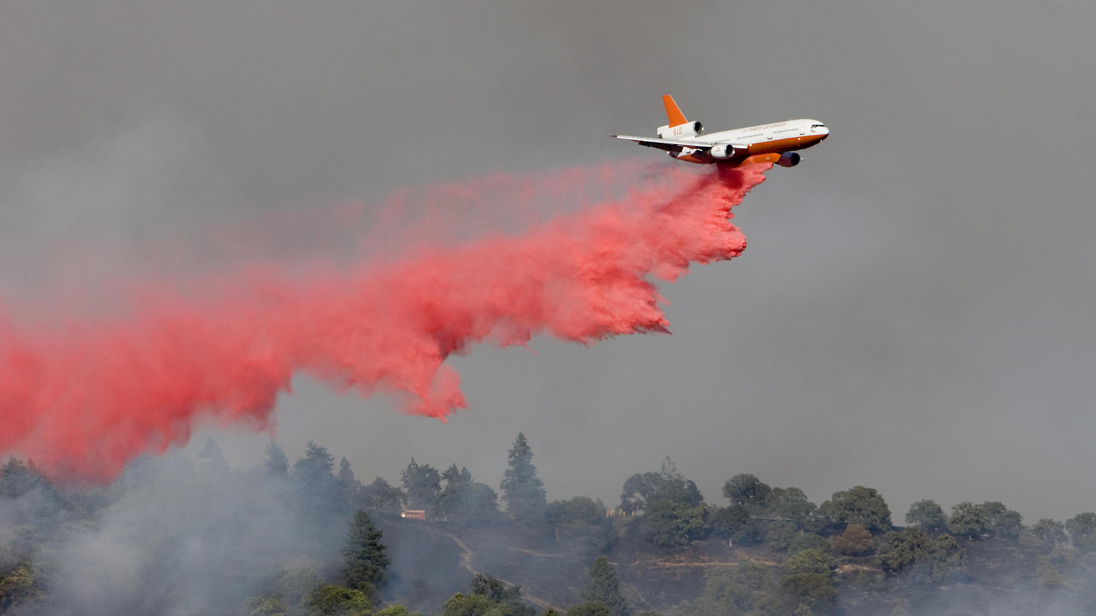 An aircraft releases red fire retardant over a forested area during wildfire suppression efforts.