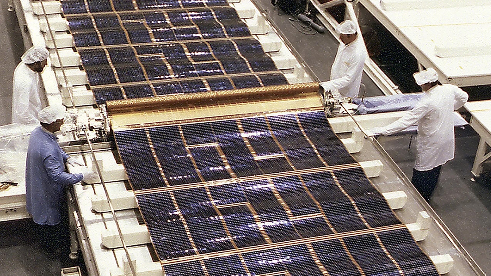 Technicians in white lab coats and hair coverings work on assembling large solar panels in a cleanroom-like environment.