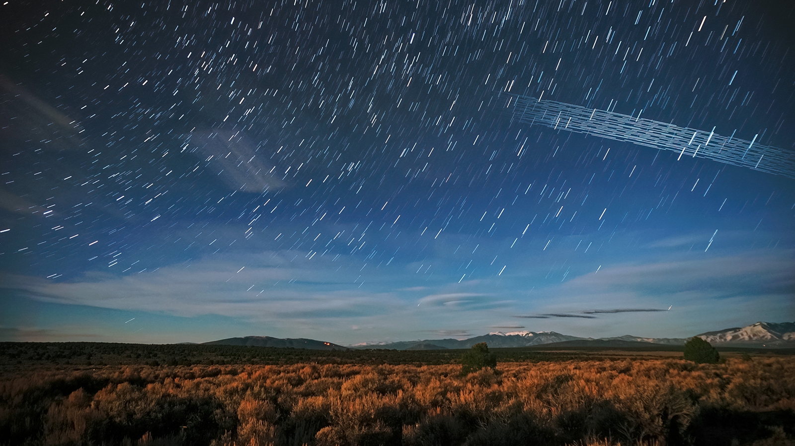 A night sky with streaks from star trails and satellites over a landscape of brush and distant mountains, under a partially cloudy sky.