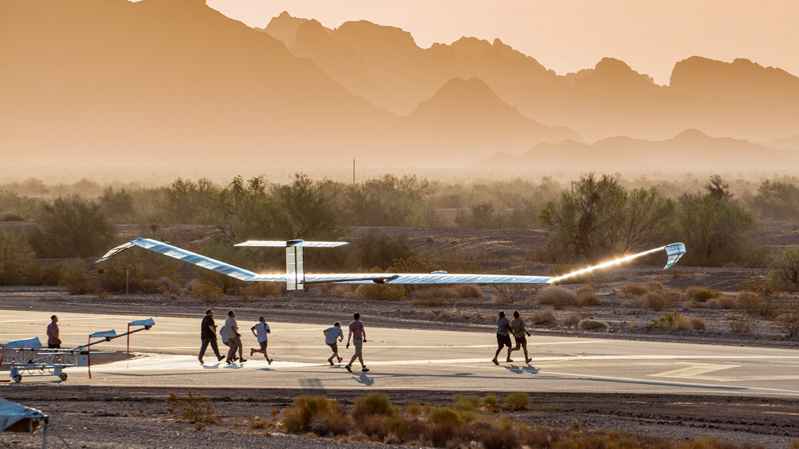 A group of people run alongside a solar-powered aircraft on a runway, set against a background of desert vegetation and mountain silhouettes at sunset.