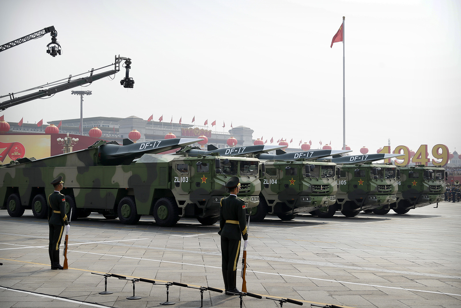 Military parade with a line of camouflaged missile launch vehicles and soldiers standing guard. A large "1949" is visible in the background.