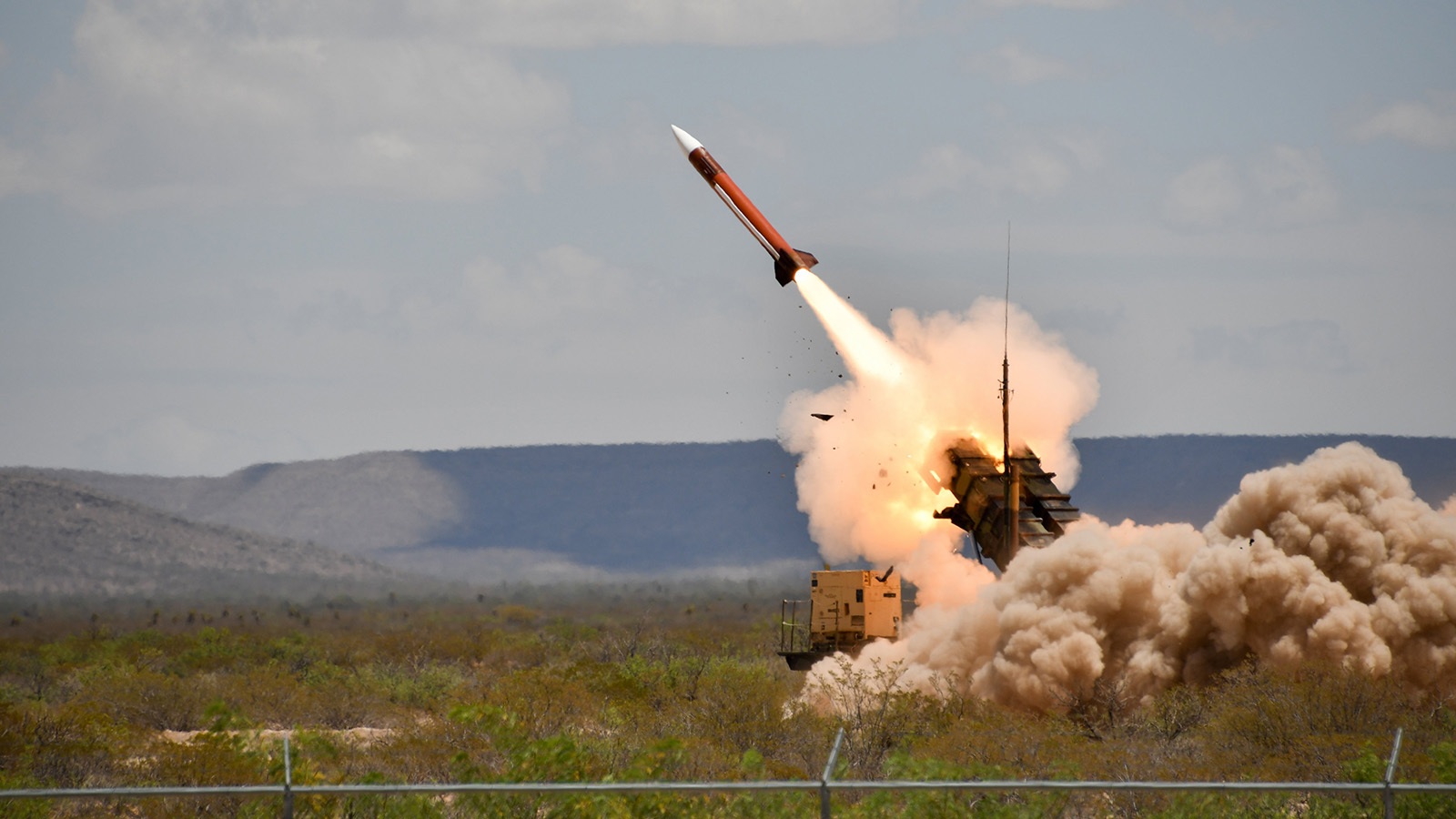 A missile is launched from a military vehicle, with smoke and flames visible, in a desert landscape under a cloudy sky.