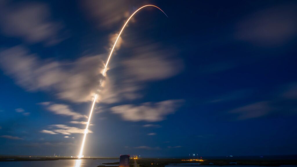 A bright arc of light from a rocket launch streaks across a cloudy night sky above a body of water, with ground lights visible below.