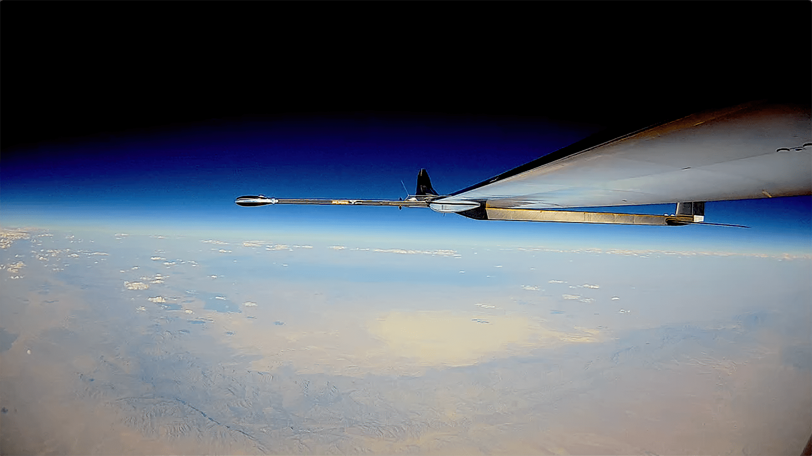 A high-altitude view from an aircraft wing, showing the curvature of the Earth and a vast landscape of deserts and mountains under a clear sky.
