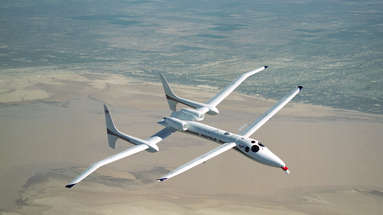A white long-winged drone with dual fuselages and tail booms flying above a desert landscape.