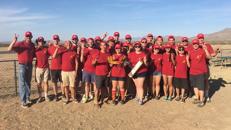 A large group of people in red shirts and hats posing cheerfully in a desert landscape, some displaying a thumbs-up gesture.