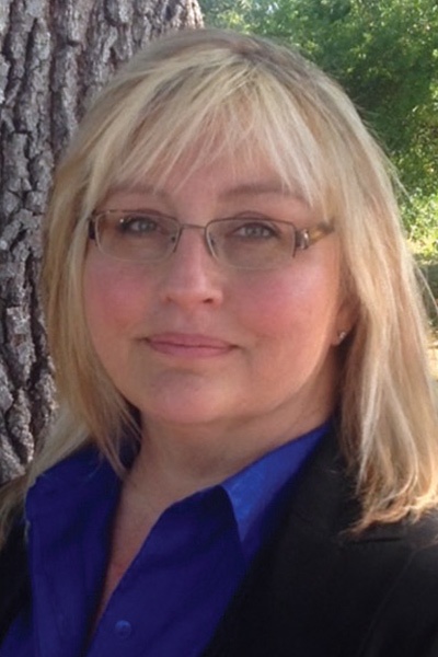 A middle-aged woman with blonde hair and glasses, wearing a blue shirt and black blazer, standing in front of a tree.