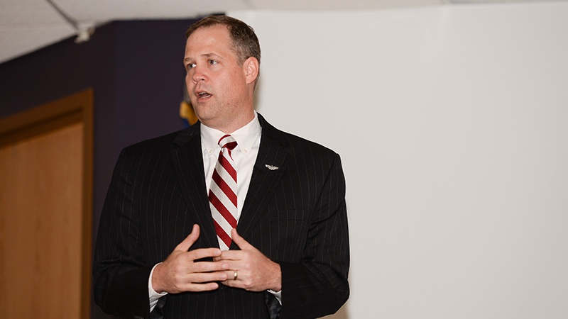 A man in a business suit with a striped tie speaks during a presentation in a room with purple walls.
