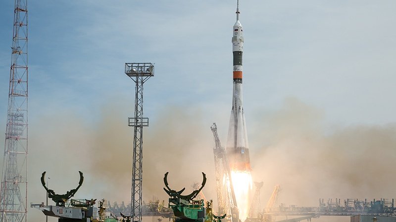 A rocket launches from a launch pad, emitting smoke and flames, with gantry towers and service structures visible against a clear sky.