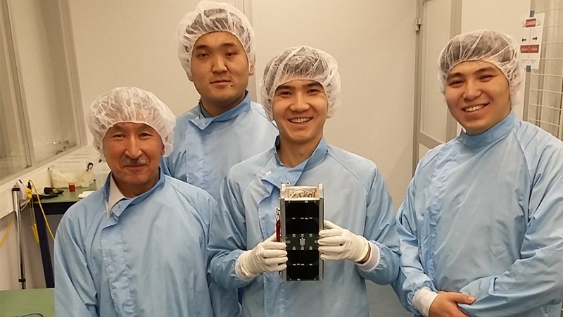 Four scientists in a cleanroom wearing blue lab coats and hairnets, smiling and holding a small satellite device.
