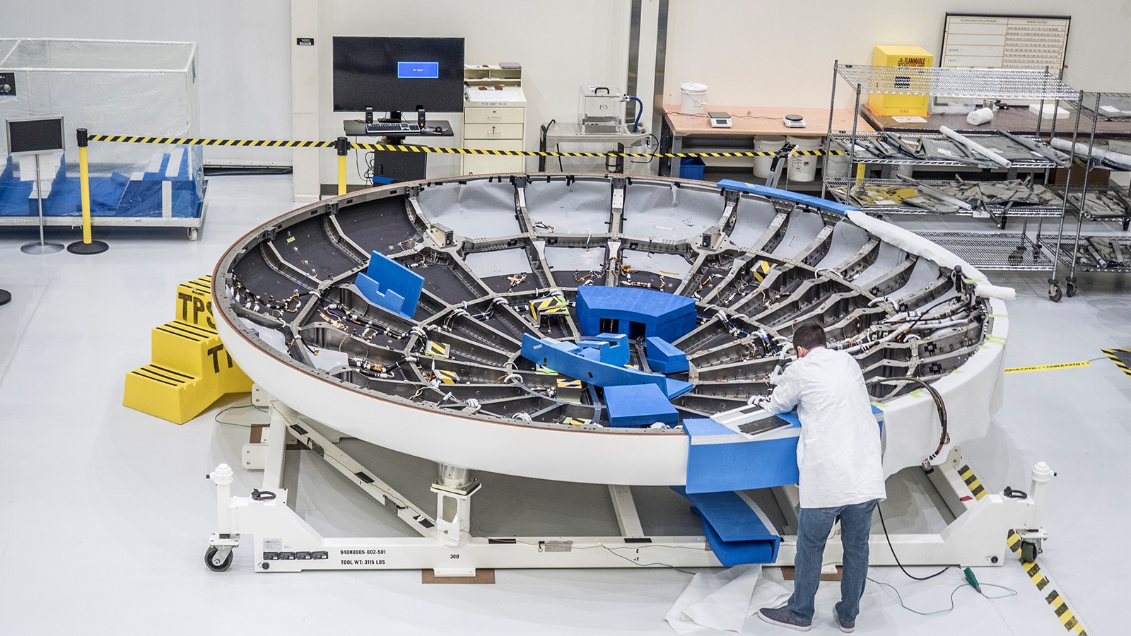 A technician in a white lab coat works on a large circular spacecraft component in a laboratory filled with equipment, shelving, and caution tape.