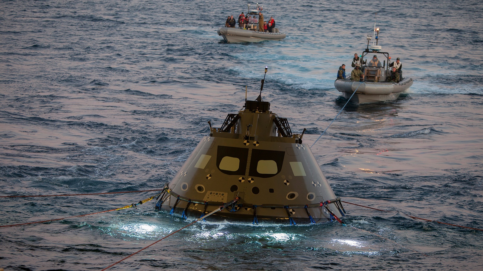 Two boats guide a space capsule floating in the ocean, equipped with lights and cables visible in the water.