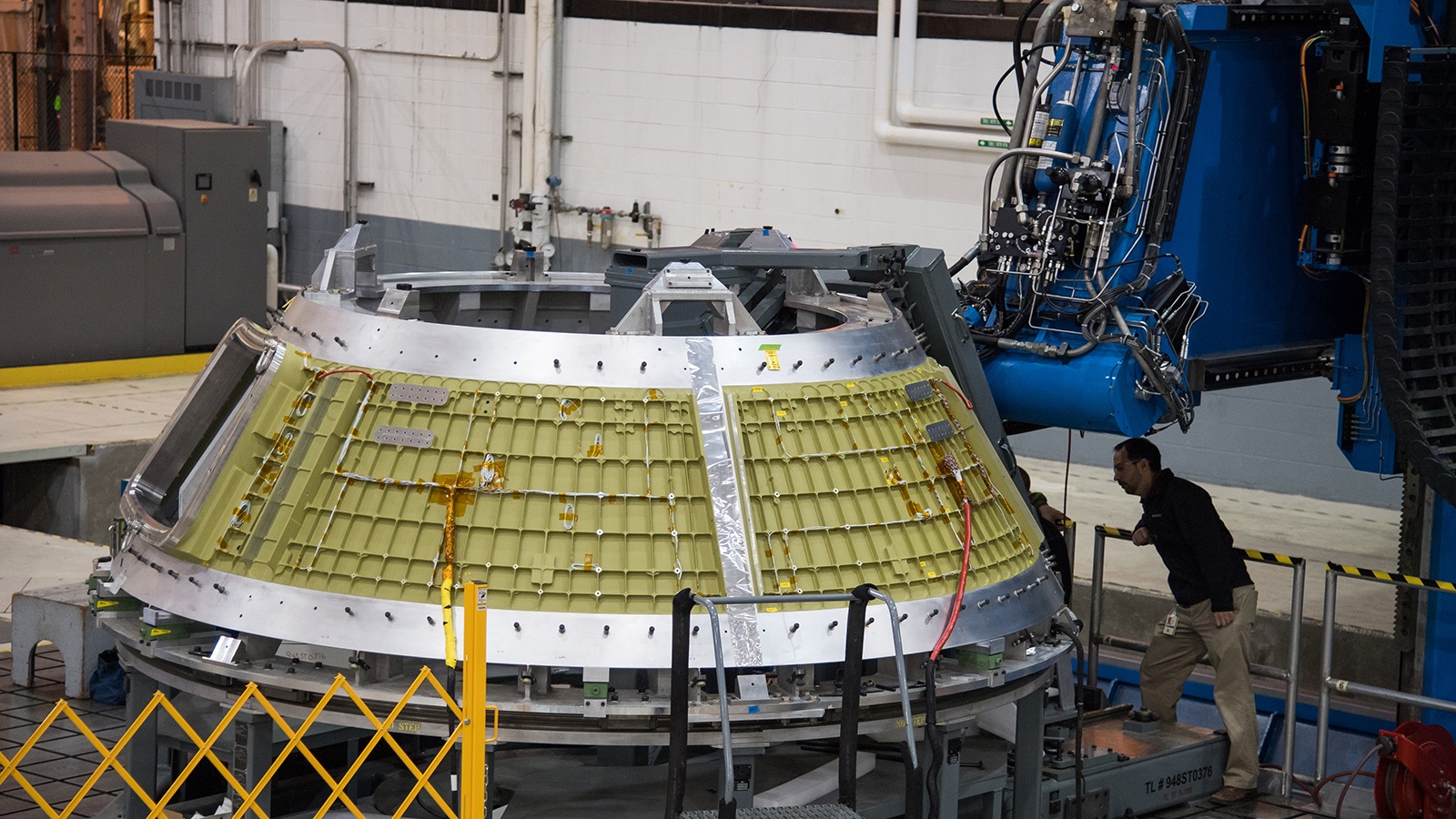 A technician inspects a partially assembled spacecraft module in an industrial setting, featuring machinery and metal structures.
