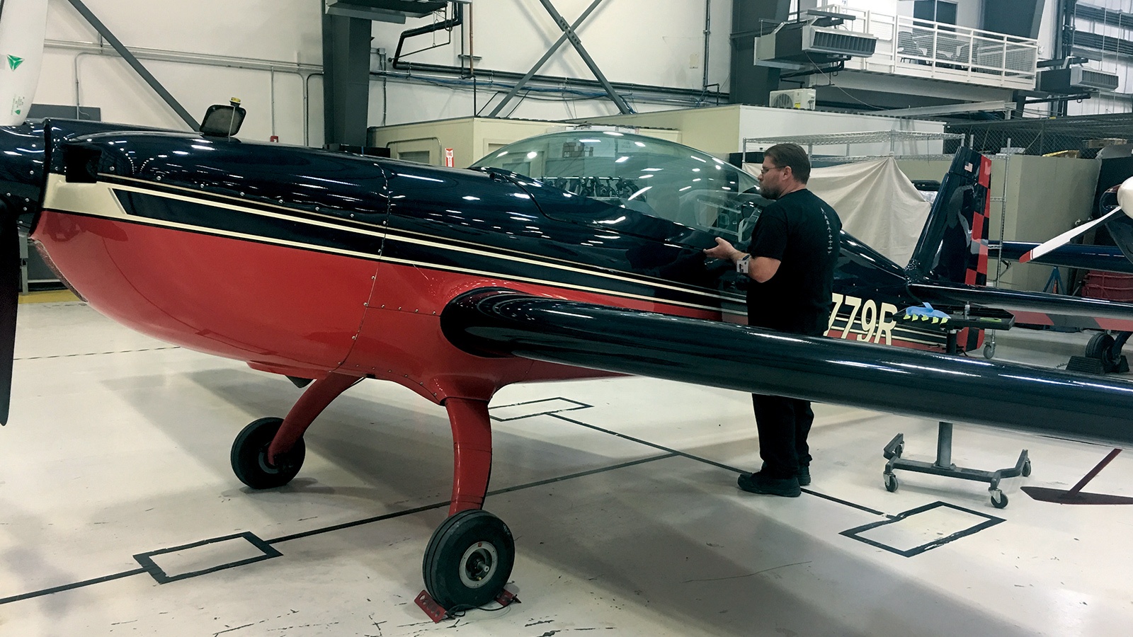 A man stands beside a small red and black airplane inside a hangar, inspecting the cockpit area. The airplane's registration number is visible on the side.