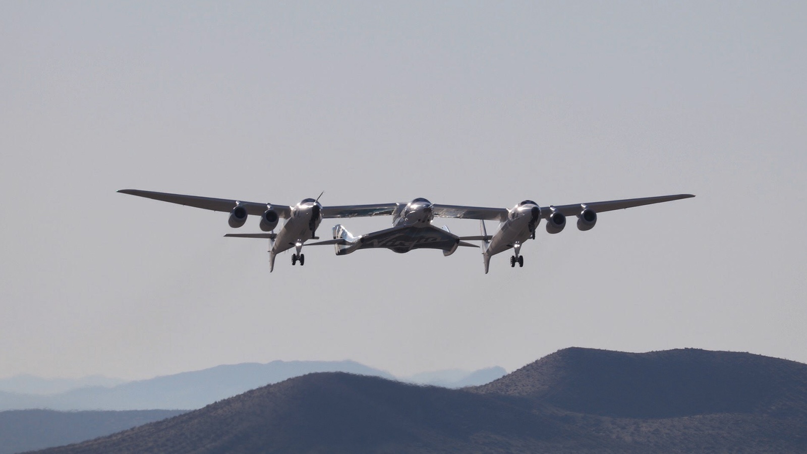 A large aircraft, consisting of two fuselages connected by a central wing, flies over a mountainous landscape under a clear sky.