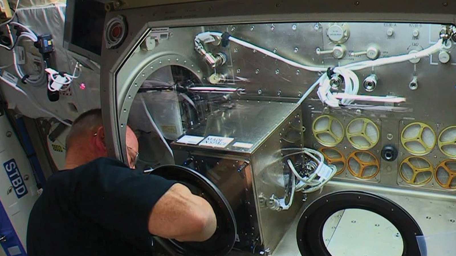 An astronaut works on equipment inside a module on the International Space Station, surrounded by cables, panels, and tools.