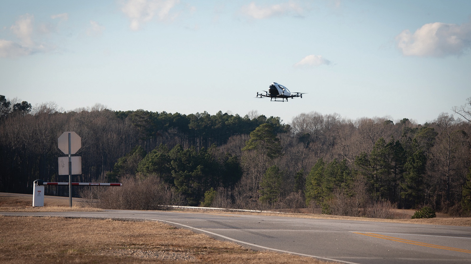 A small drone flies above a rural road, with trees lining the background and a few clouds in the sky. A stop sign and gate are visible on the left side of the road.