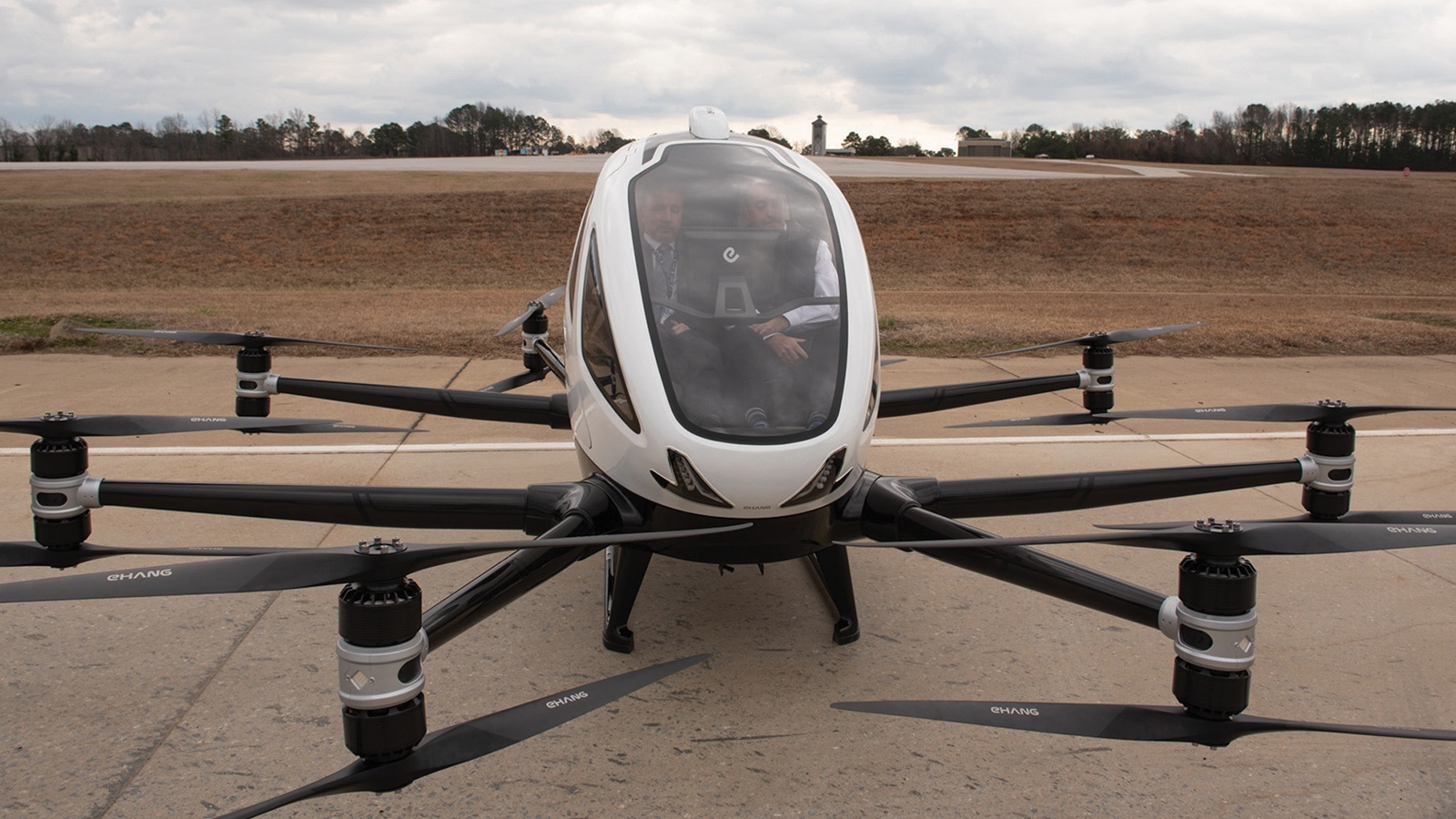 A white multicopter aircraft with eight rotors is stationed on a concrete surface. Two people are seated inside the cockpit, preparing for a flight. A runway and some trees are visible in the background.