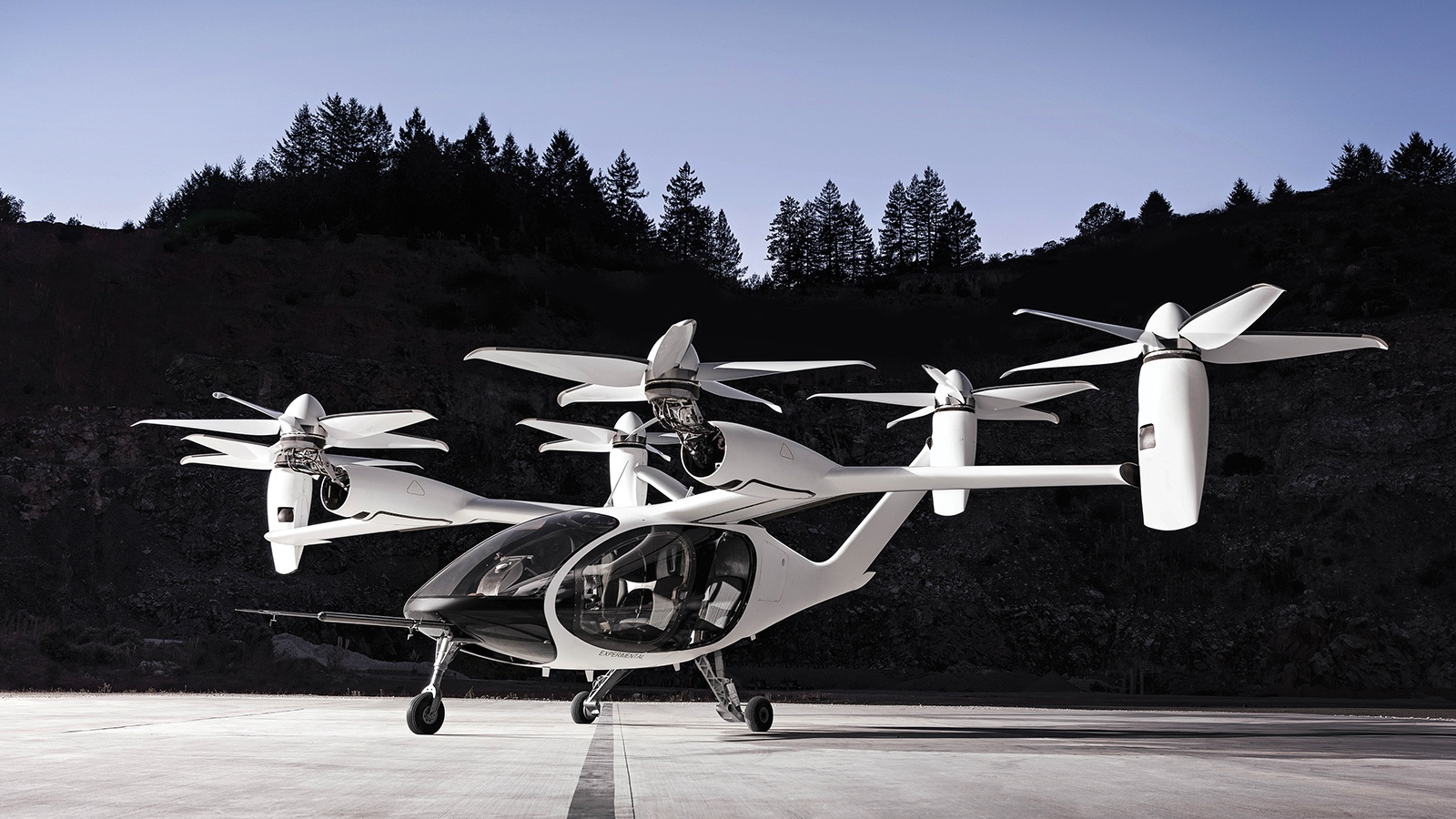 A sleek white electric vertical takeoff and landing aircraft (eVTOL) is parked on a flat surface with forested hills in the background. It has six rotors and a compact, futuristic design.
