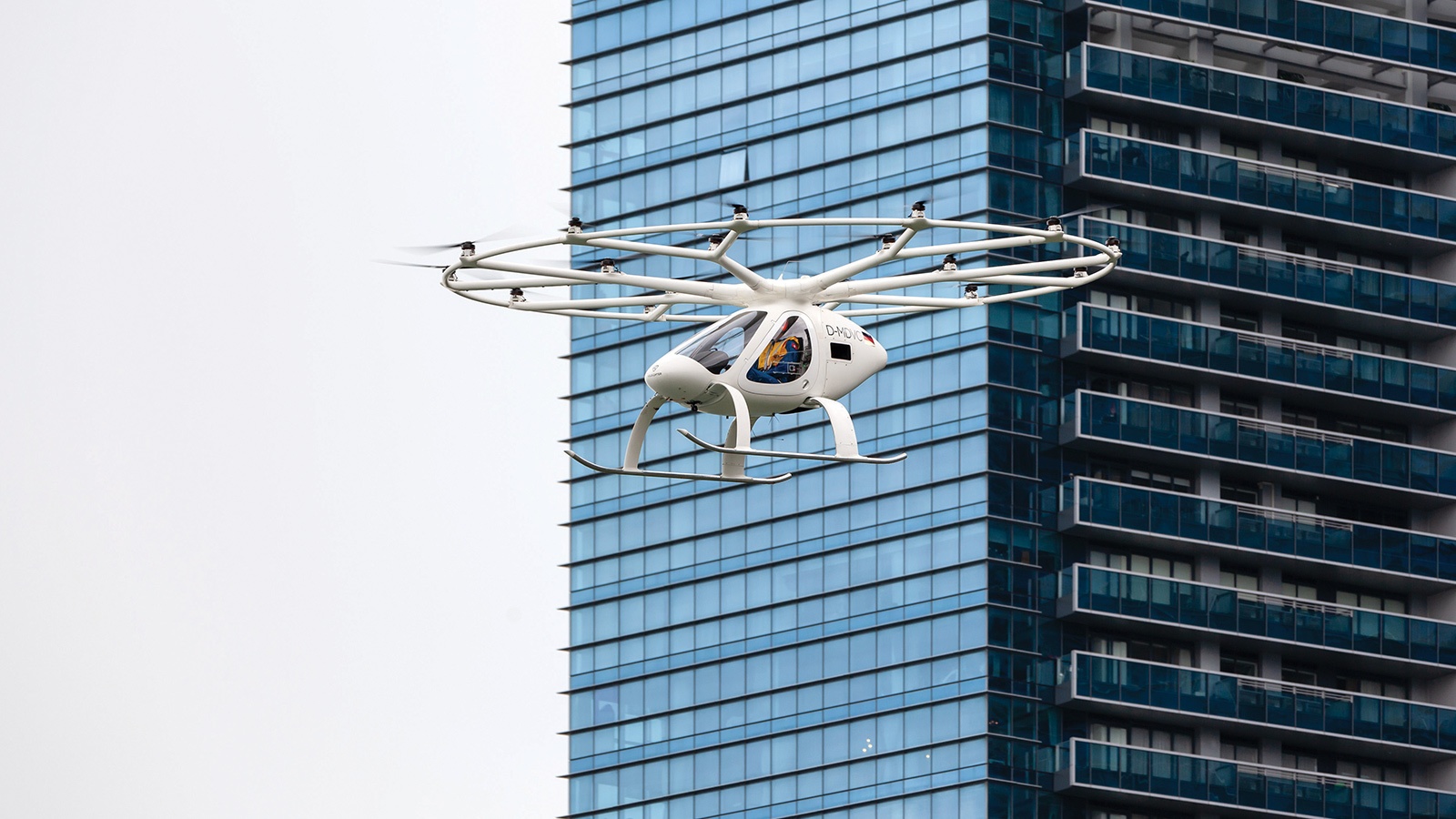 A white multicopter drone is flying in front of a tall, blue-glass building on an overcast day.