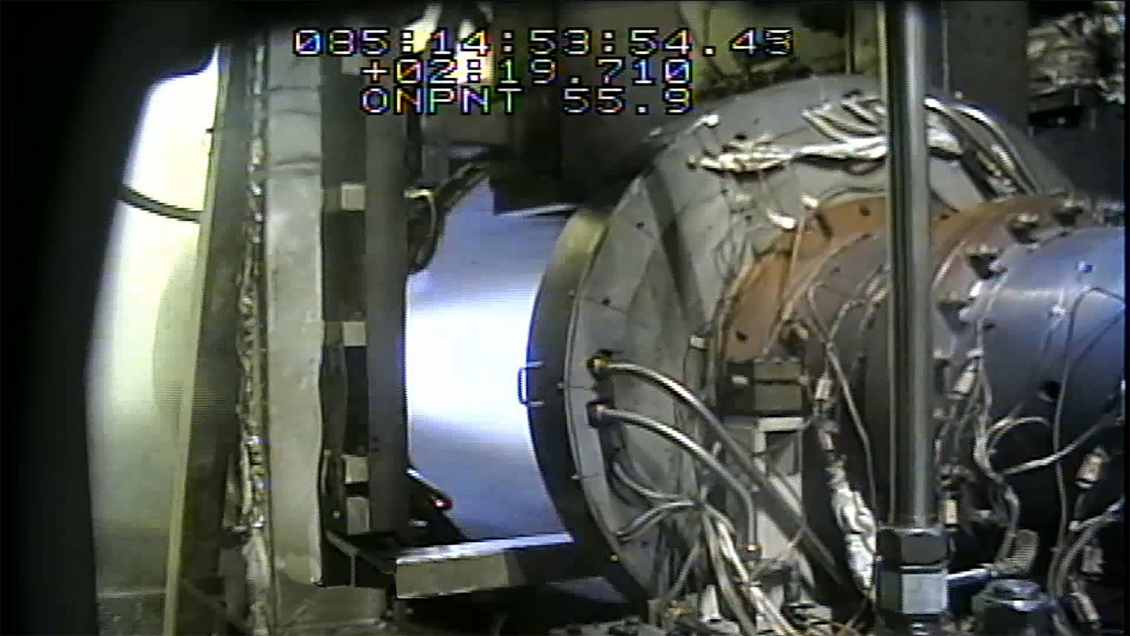 A view inside an industrial facility showing machinery with numerous cables and wires attached. The image includes timestamp and measurement data overlayed on the top part.