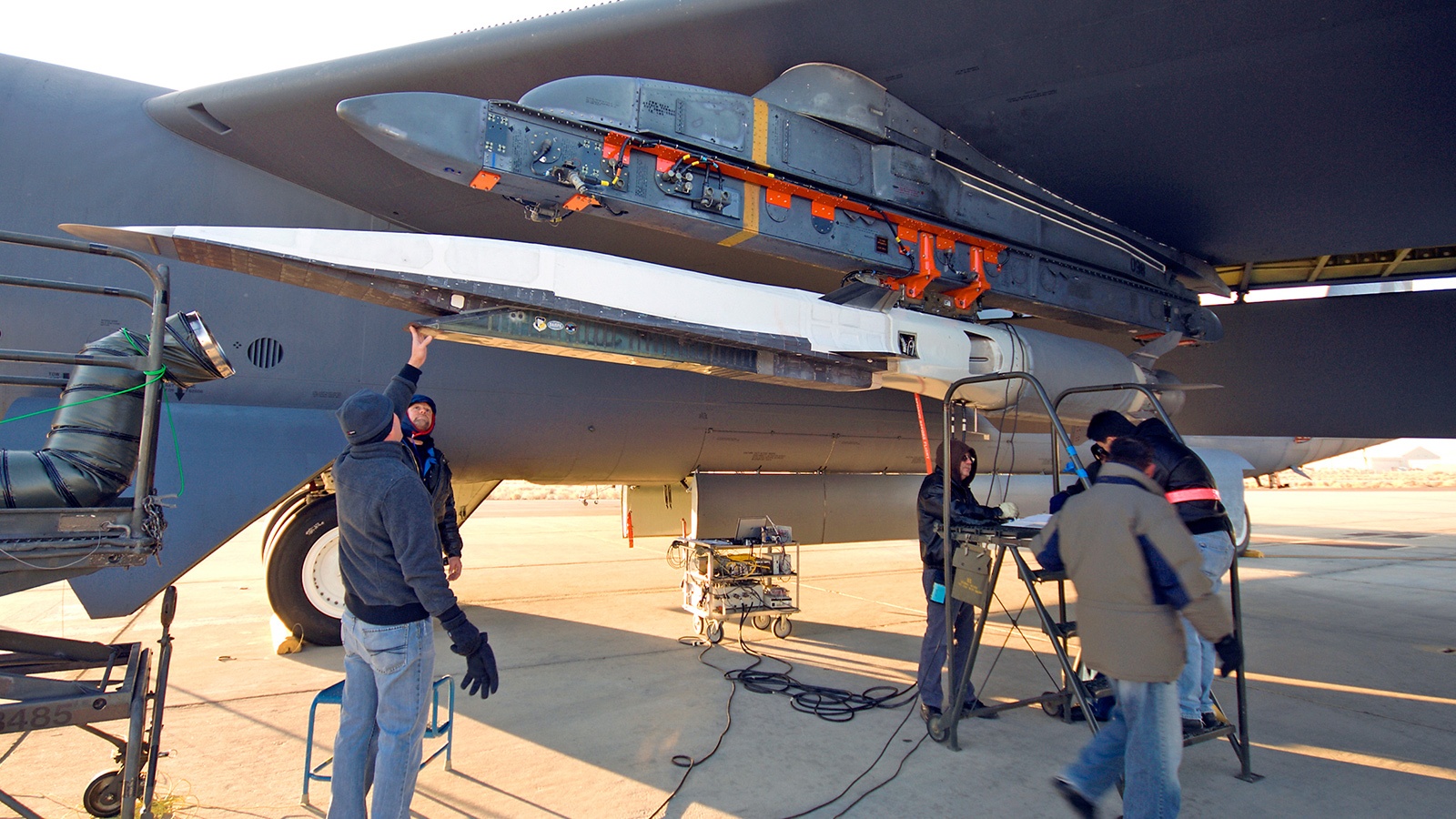 Engineers work on attaching a missile under the wing of a large military aircraft on an outdoor airfield. There are tools and equipment on the ground, and the aircraft's landing gear is visible.