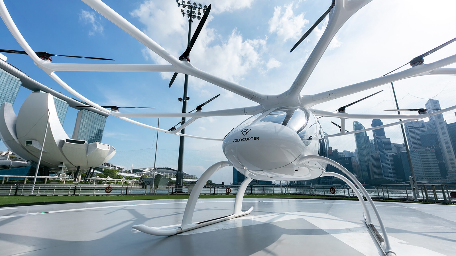 A white Volocopter air taxi is parked on a helipad with a city skyline and a blue sky with clouds in the background.
