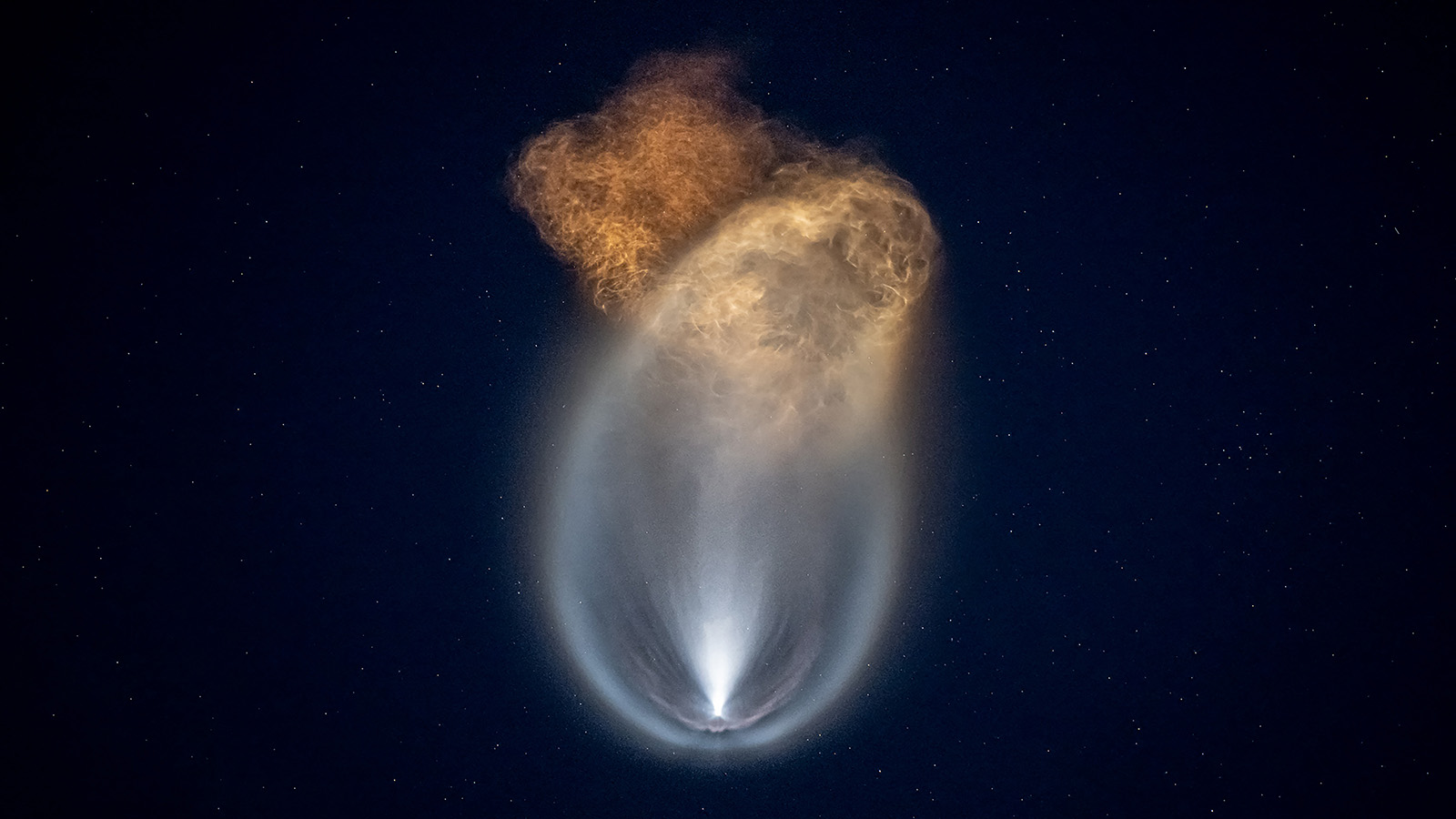 A rocket or spacecraft leaving a glowing trail against a starry night sky, with a bright plume transitioning from white to reddish-orange at the top.