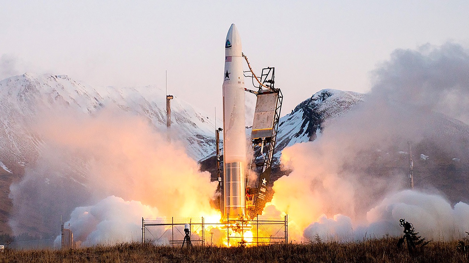 A rocket launches from a mountainous area with flames and smoke billowing from its engines, against a backdrop of snow-covered peaks.