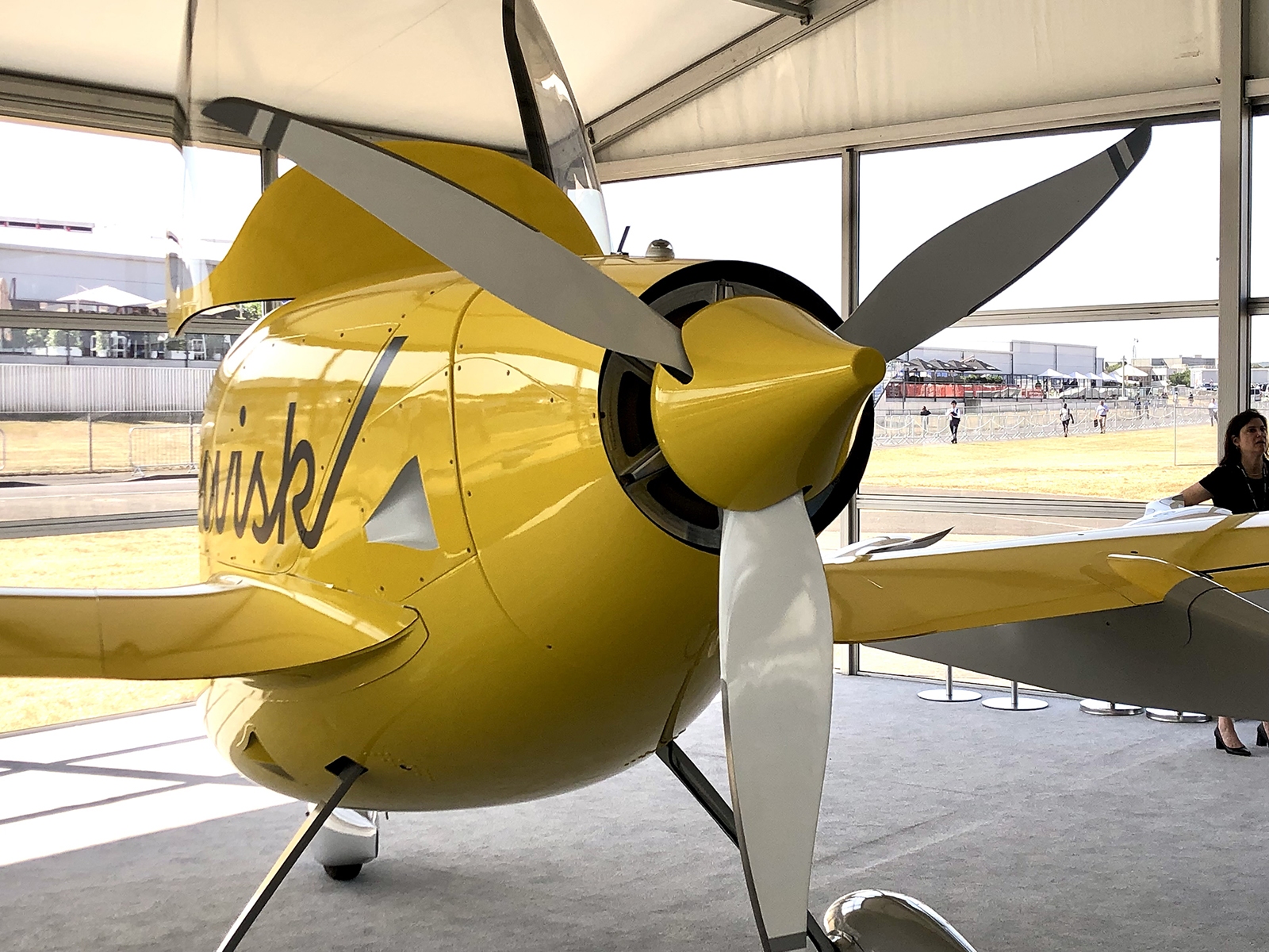 Yellow, single-engine aircraft on display inside a tent at an airshow, showcasing its propeller and front section with a partial view of the aircraft's name on the side.