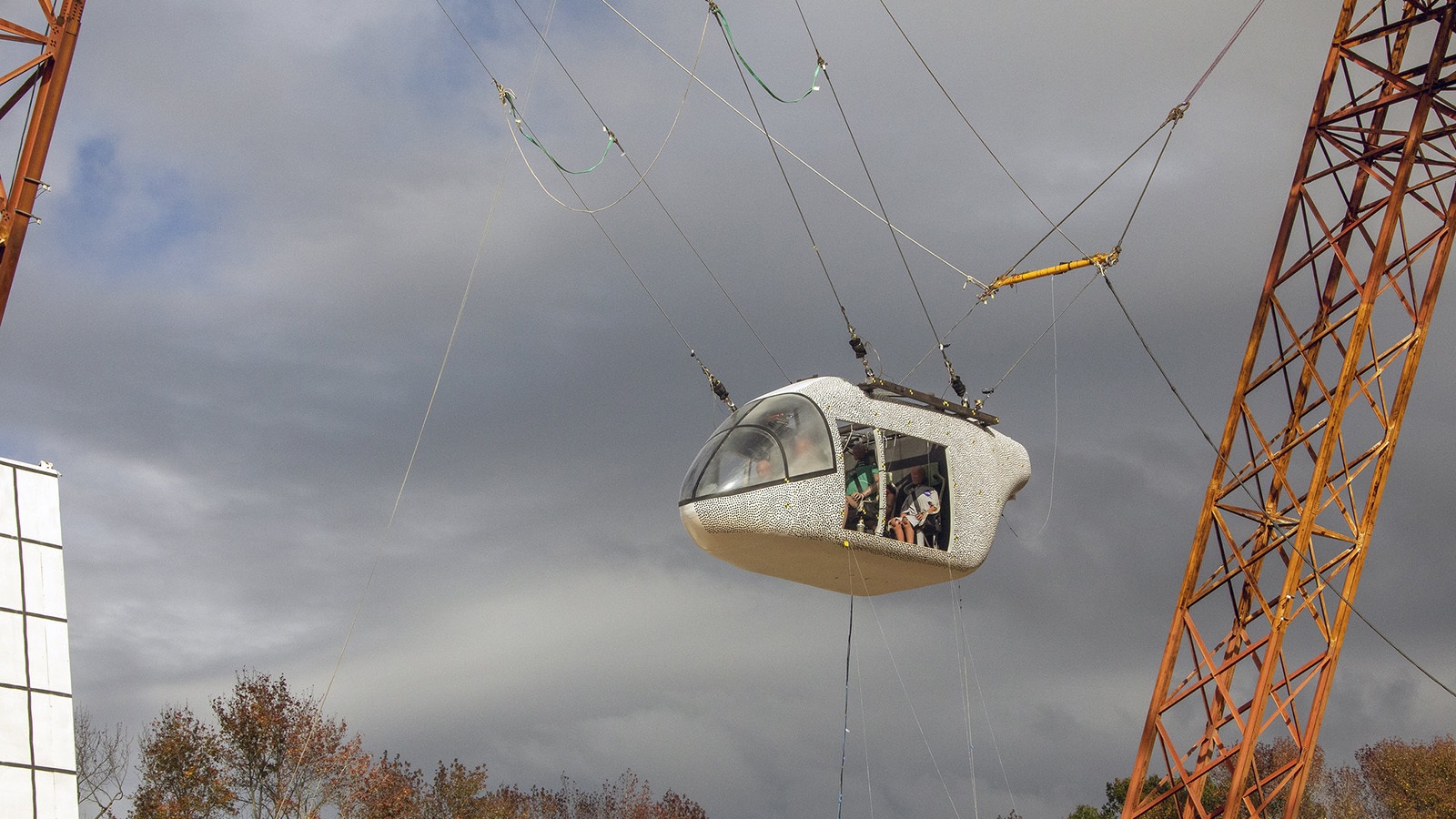 A small, white, pod-like capsule suspended in mid-air by cables attached to a structure, with people visible inside it. The sky is overcast, and autumn trees are in the background.