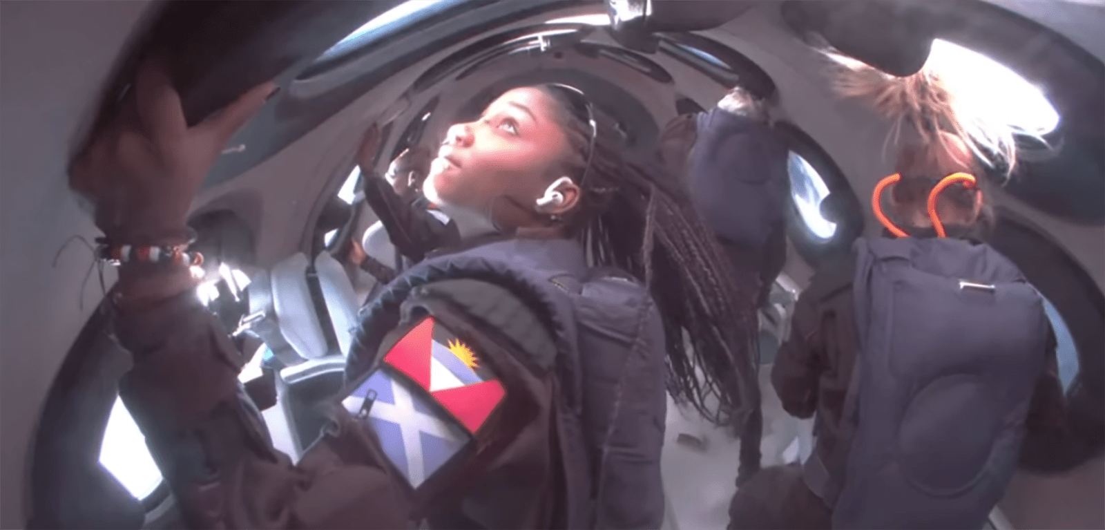 Astronauts experience floating inside a spacecraft, a woman looks out a window, wearing a suit displaying a colorful patch with a flag.