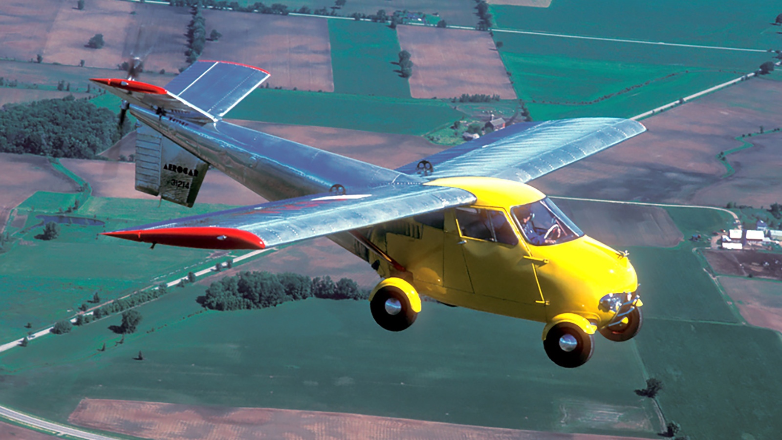 A small yellow and silver car-plane hybrid with extended wings is flying over a patchwork of agricultural fields.
