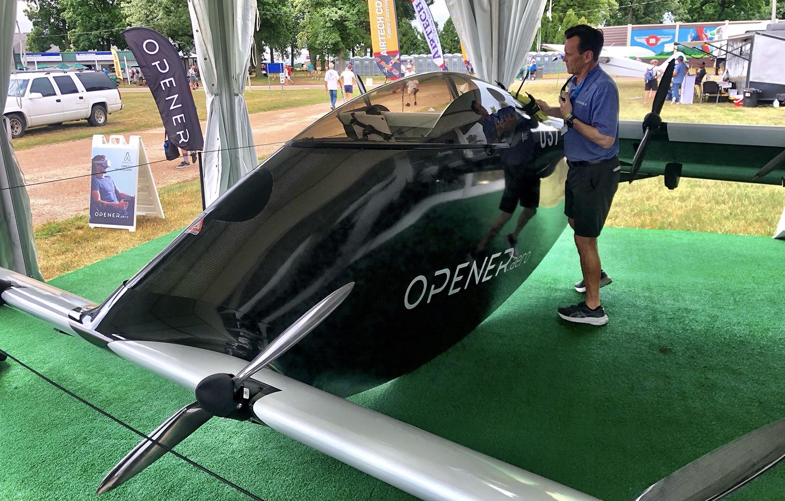 A man stands next to an Opener BlackFly aircraft showcased under a tent at an outdoor event. The aircraft has a sleek black body and a large propeller at the front.