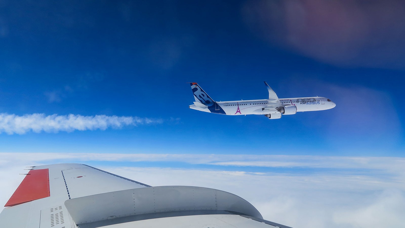 An airplane in flight viewed from the window of another aircraft, with a blue sky and clouds in the background.