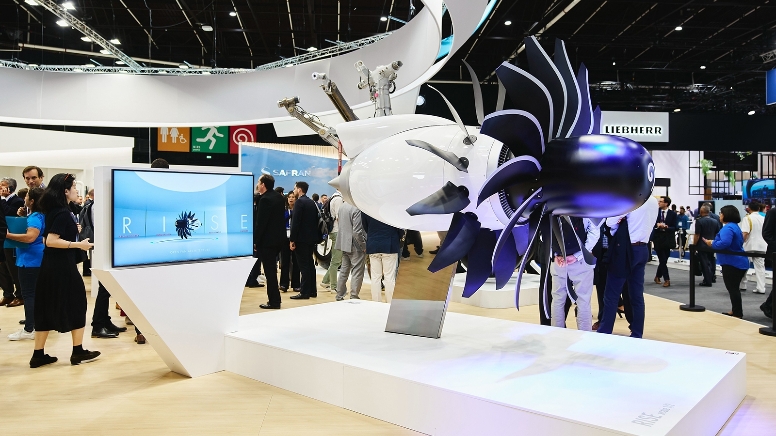 A turbine engine model is displayed on a white platform at an exhibition, with people gathered around and viewing it. A screen on the left displays related information about the engine.