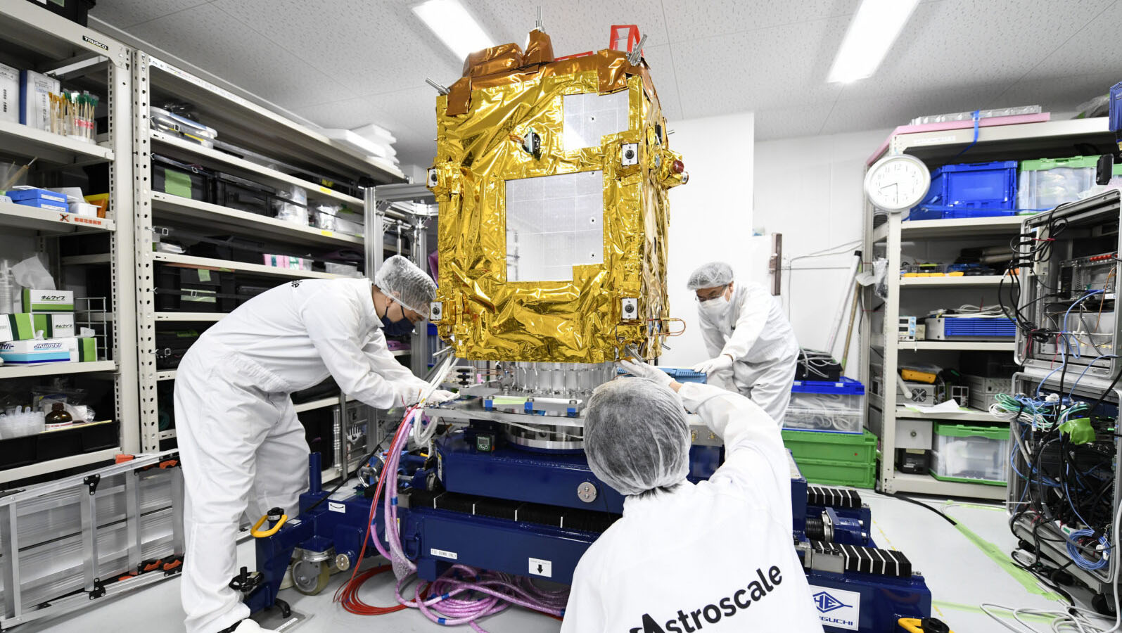Workers in white protective suits assemble a satellite covered in gold foil within a laboratory setting equipped with various tools and equipment.