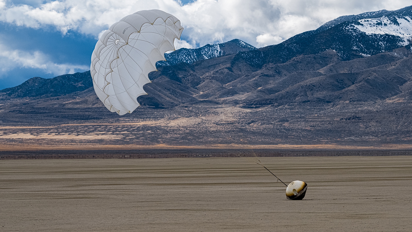 A parachute and capsule on a flat desert landscape with mountains in the background under a cloudy sky.