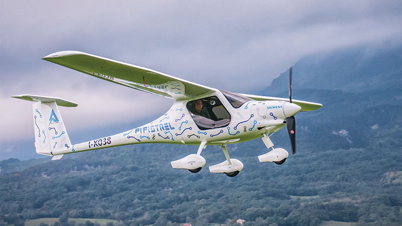 A small green and white electric airplane in flight against a cloudy mountainous backdrop.