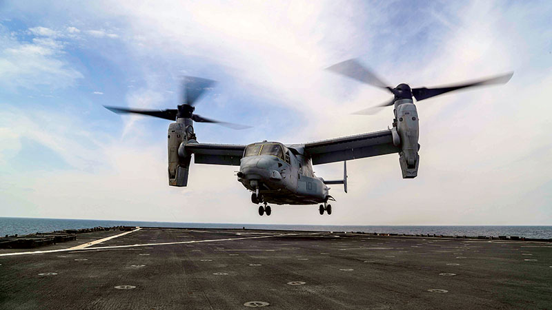 A v-22 osprey tiltrotor aircraft taking off from a ship's flight deck, with its rotors in vertical position under a cloudy sky.