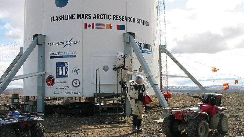 A person in a spacesuit stands outside the flashline mars arctic research station, surrounded by scientific equipment and atvs, in a barren landscape.