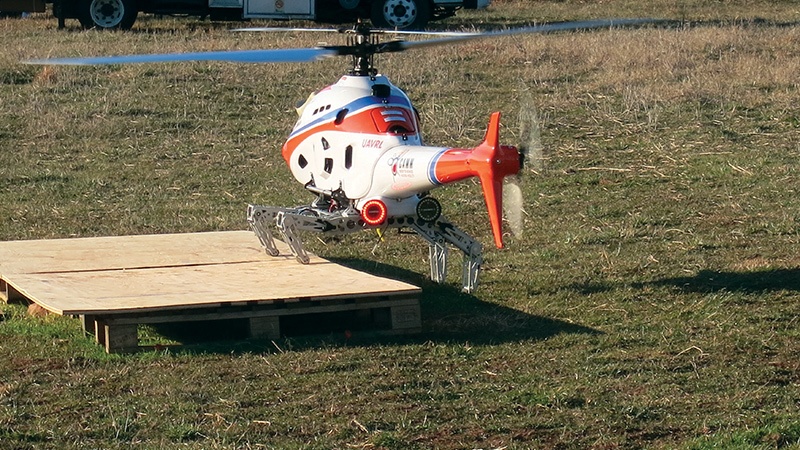 A remote-controlled helicopter with a white, blue, and red color scheme hovers above a wooden ramp in a grassy field.