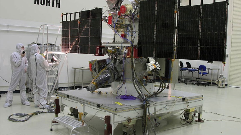 Technicians in clean suits work on a large satellite with extended solar panels in a clean room facility.